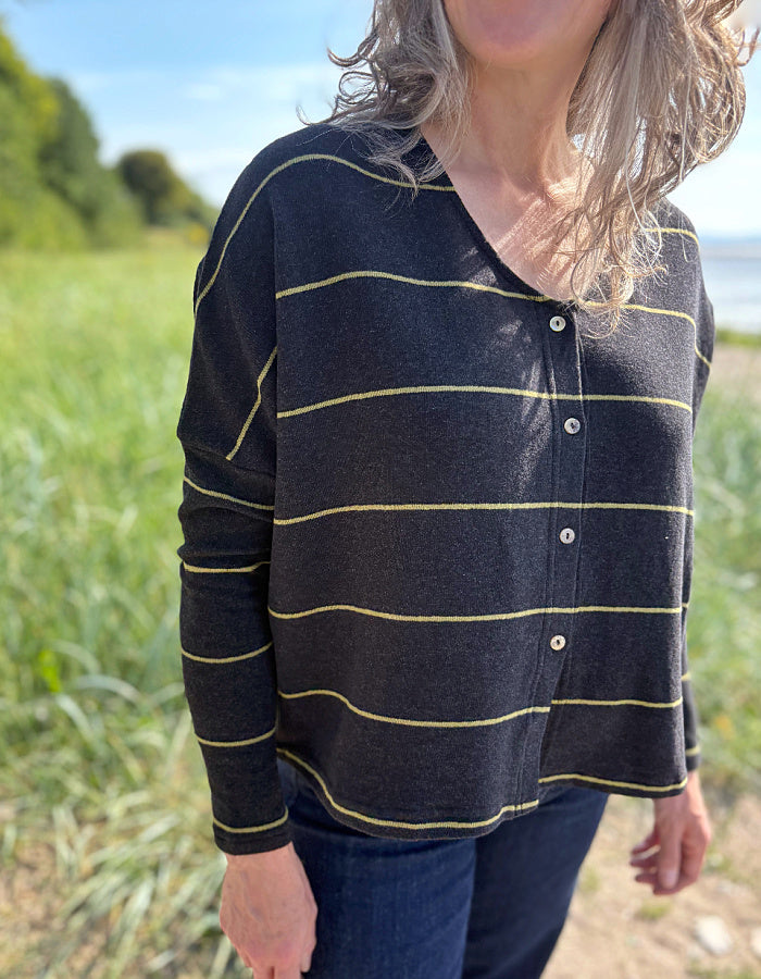 Woman wearing charcoal and olive pin stripe cardigan and sunglasses, standing in a grassy field with a body of water in the background.