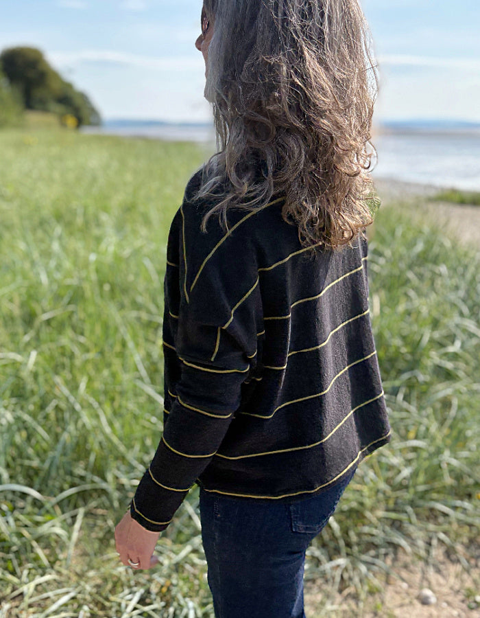 Woman wearing charcoal and olive pin stripe cardigan and sunglasses, standing in a grassy field with a body of water in the background.