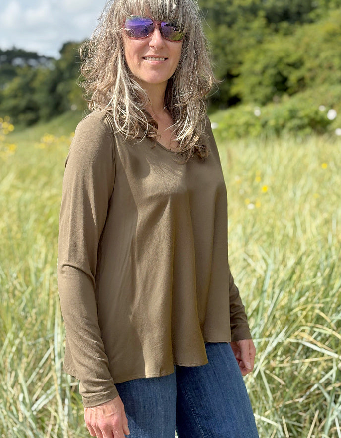 Woman wearing a brown long-sleeve top and sunglasses standing in a field with greenery in the background.
