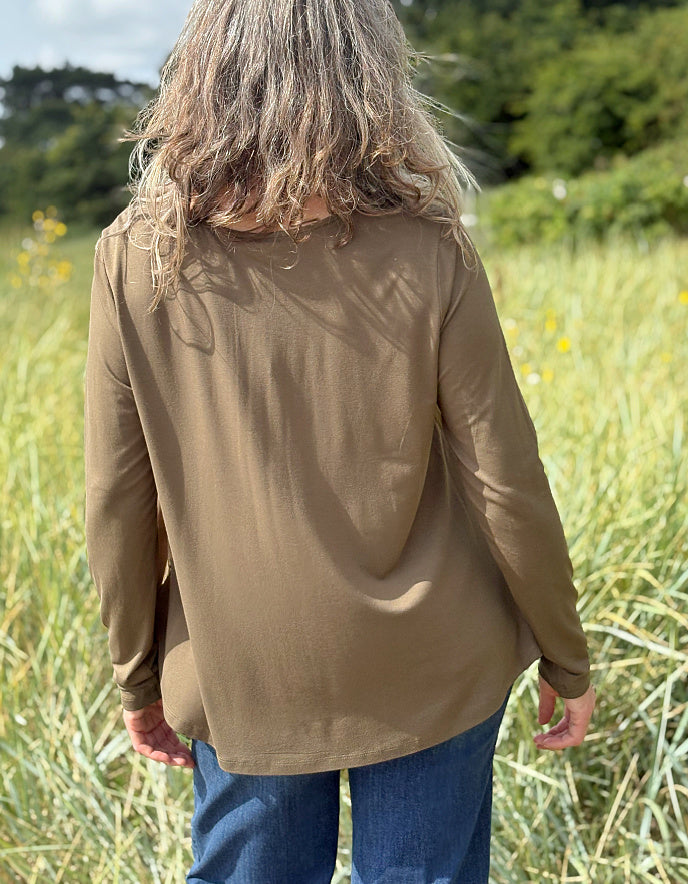 Person wearing a brown jacket standing in a field with greenery and yellow flowers in the background