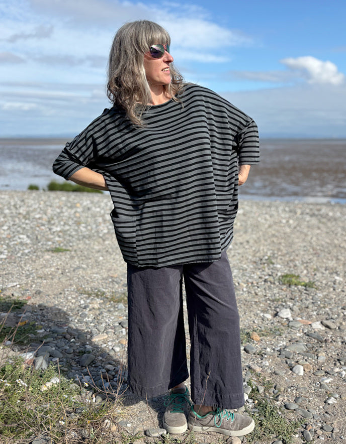 Woman standing on a pebbly beach wearing a striped black and grey oversized sweatshirt top  and sunglasses.