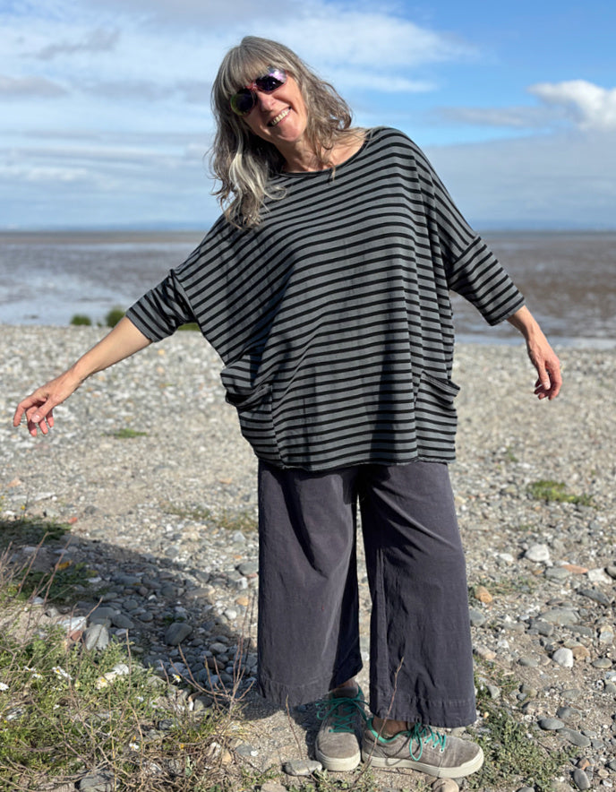 Woman standing on a pebbly beach wearing a striped black and grey oversized sweatshirt top  and sunglasses.