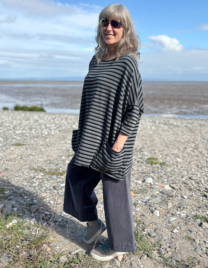 Woman standing on a pebbly beach wearing a striped black and grey oversized sweatshirt top  and sunglasses.