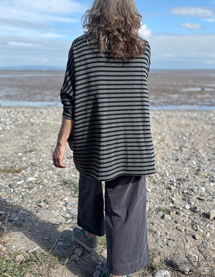Woman standing on a pebbly beach wearing a striped black and grey oversized sweatshirt top  and sunglasses.