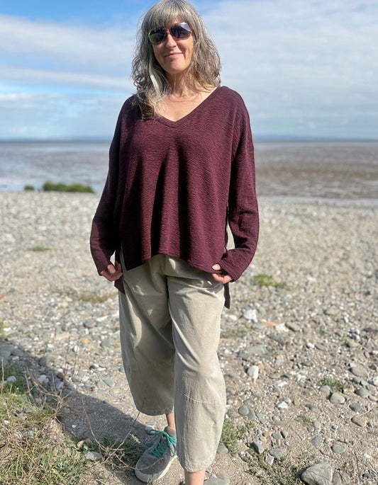 Woman standing on a pebbly beach wearing a maroon sweater and beige pants.