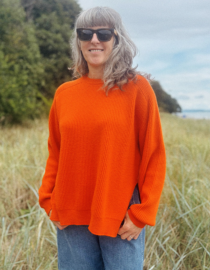 A woman with gray hair and sunglasses wearing a bright orange ribbed knit sweater and blue jeans, standing in a grassy outdoor field.