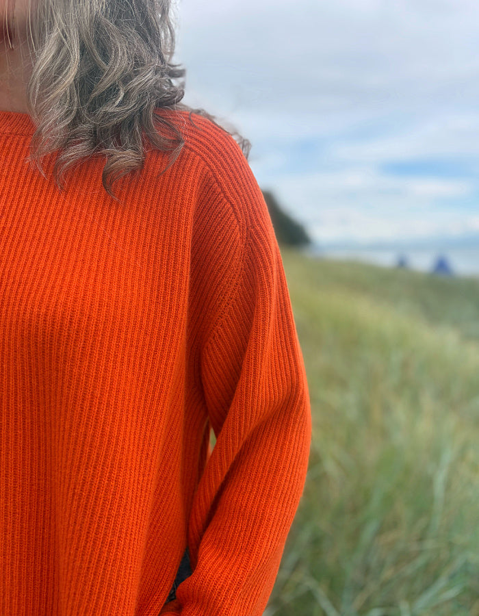 A close-up of a person wearing a bright orange ribbed knit sweater outdoors in a grassy field with a cloudy sky in the background.