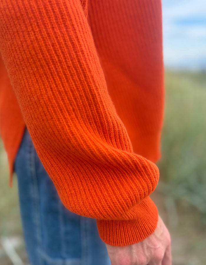 Close-up of a person wearing a bright orange ribbed knit sweater, showing the sleeve cuff and part of the blue jeans, with a blurred outdoor background.