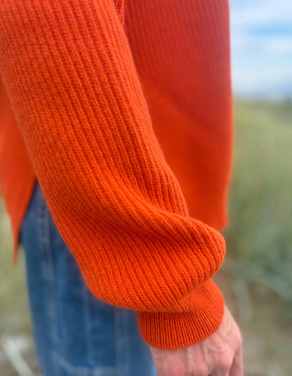 Close-up of a person wearing a bright orange ribbed knit sweater, showing the sleeve cuff and part of the blue jeans, with a blurred outdoor background.