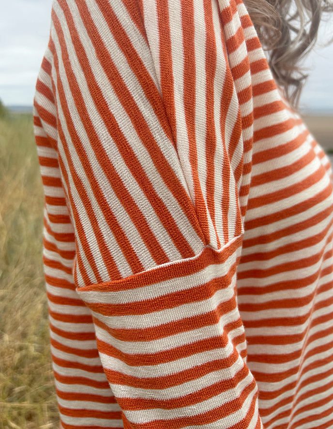 Close-up of a person wearing a knit top with orange and off-white horizontal stripes, photographed outdoors with a blurred grassy field and sky in the background.