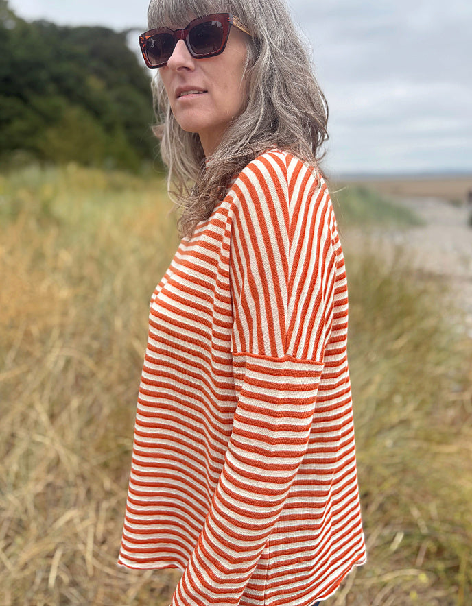 A woman with gray hair and sunglasses wearing a rust-orange and white striped knit top, standing outdoors in a grassy field.