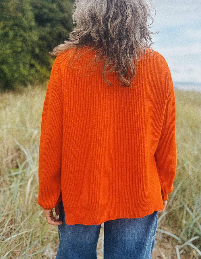 A person standing outdoors in a grassy field, viewed from behind, wearing a bright orange ribbed knit sweater and blue jeans.