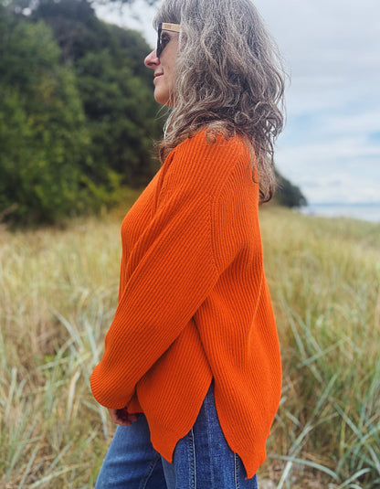 A woman wearing a bright orange ribbed knit sweater and blue jeans stands outdoors in a grassy field with trees and a cloudy sky in the background.