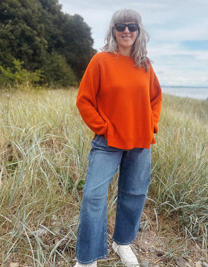 A woman wearing a bright orange knit sweater, blue wide-leg jeans, and white sneakers stands outdoors in a grassy field near trees and water, wearing dark sunglasses.
