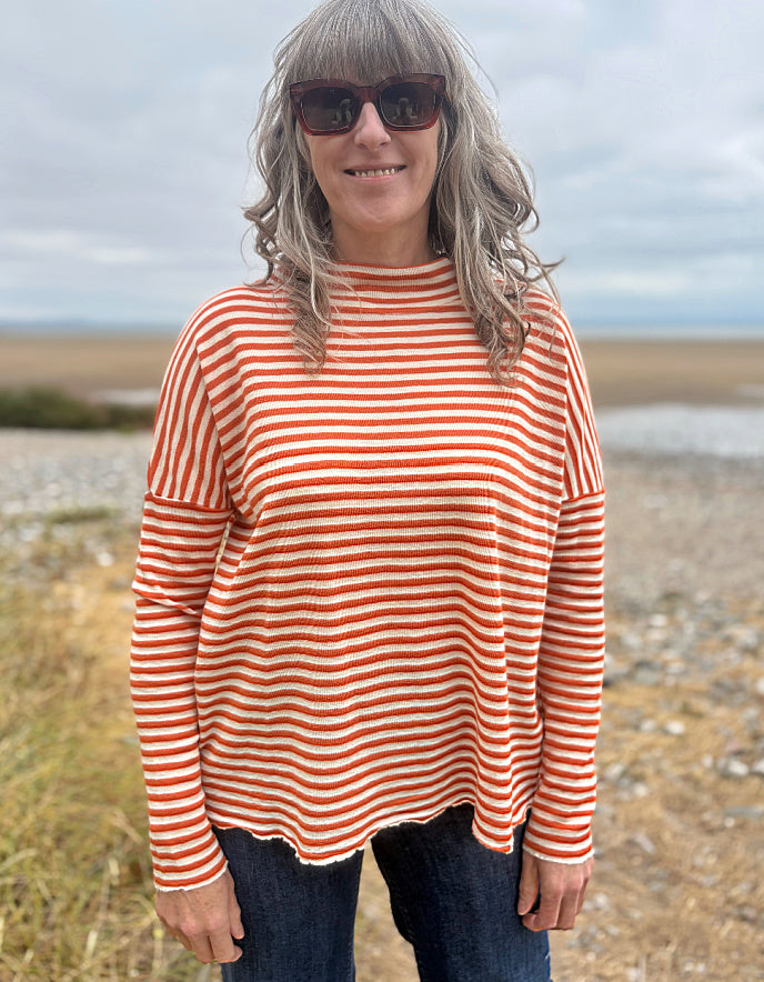 A woman with gray hair and sunglasses wearing a rust-orange and white striped long-sleeve top, standing outdoors on a beach or pebble shore.