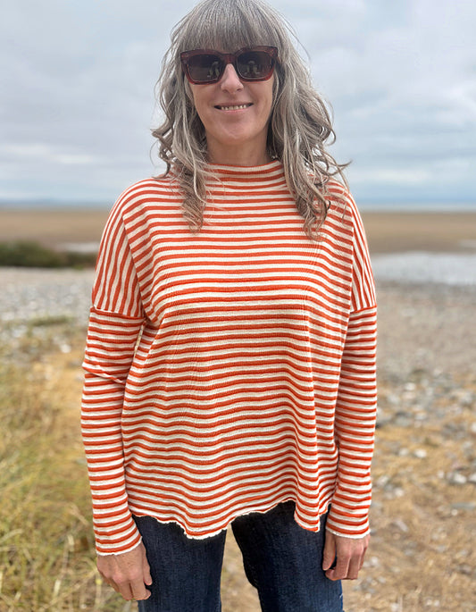 A woman with gray hair and sunglasses wearing a rust-orange and white striped long-sleeve top, standing outdoors on a beach or pebble shore.