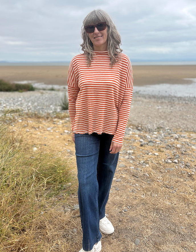 A woman standing on a rocky beach wearing a long-sleeve orange and white striped knit top, dark blue jeans, and white sneakers.