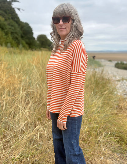 A woman with gray hair and sunglasses stands in a grassy outdoor field wearing a long-sleeve orange and white striped top with blue jeans.