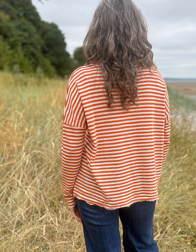 A woman viewed from behind wearing a rust-orange and white horizontally striped long-sleeve top and dark blue jeans, standing in a grassy outdoor field under an overcast sky.