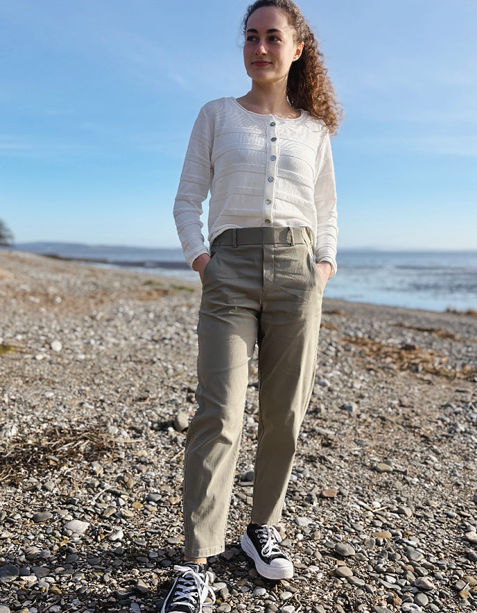 Woman standing on a pebbly beach wearing a light-colored cardigan and beige pants.