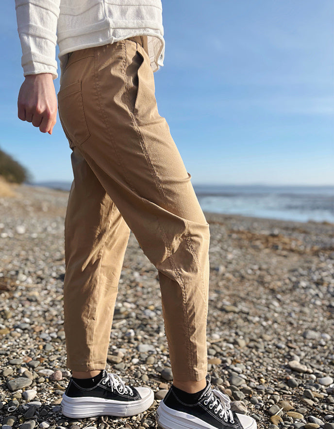Person wearing beige pants and black sneakers on a pebbly beach with ocean in the background