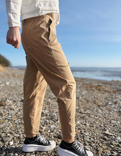 Person wearing beige pants and black sneakers on a pebbly beach with ocean in the background