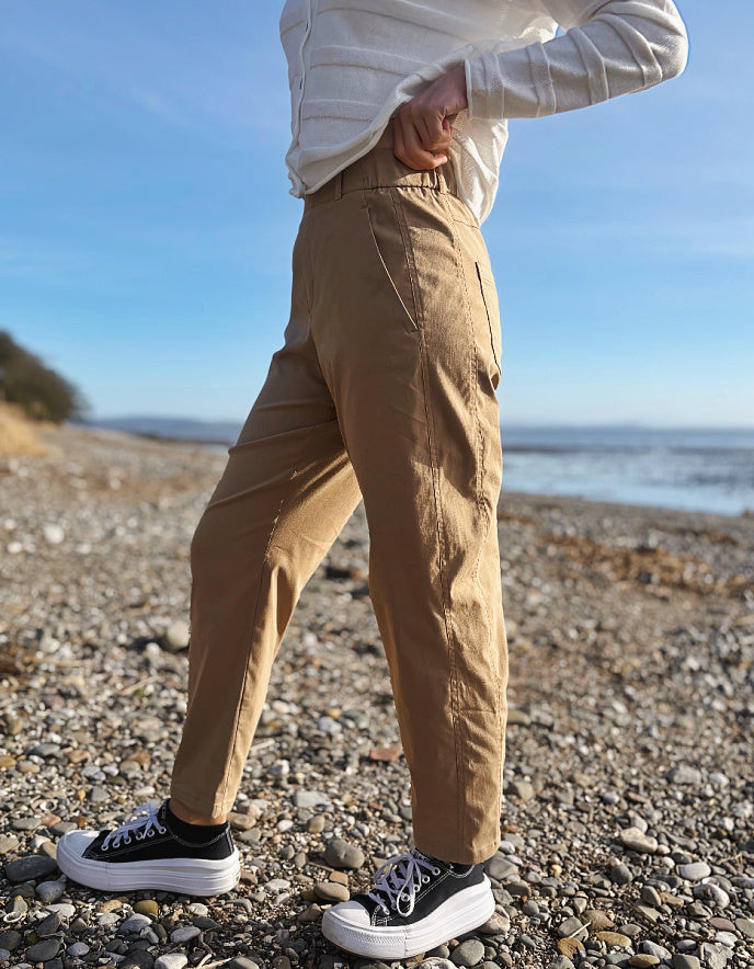 Person wearing beige pants and black sneakers standing on a pebbly beach with ocean in the background
