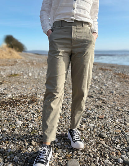 Person wearing beige pants and black sneakers standing on a pebbly beach with ocean in the background