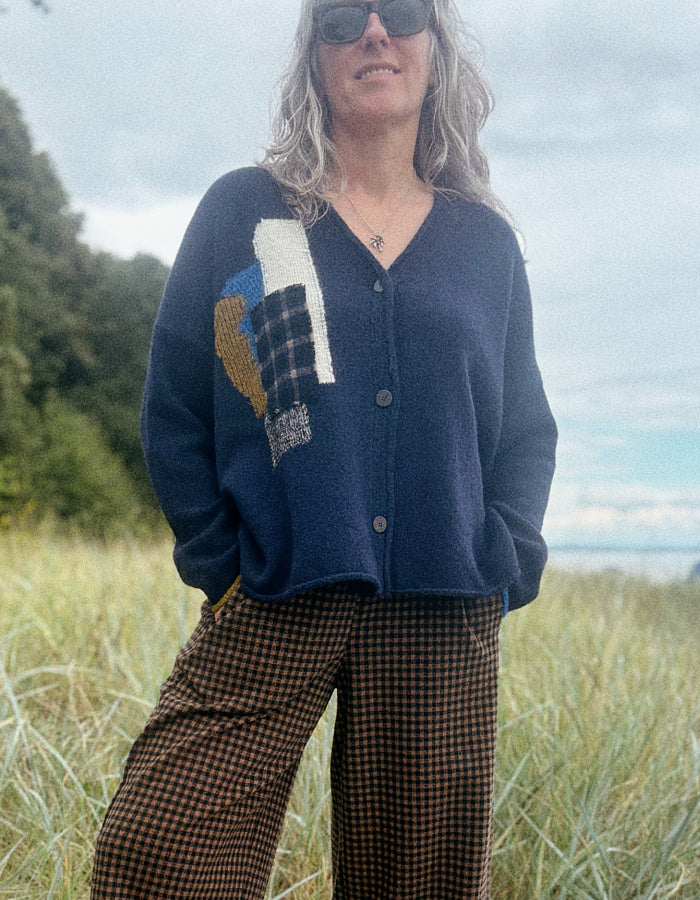 A woman wearing a navy blue knit cardigan with a colorful patchwork design on the front and brown checkered pants, standing in a grassy outdoor field under an overcast sky.