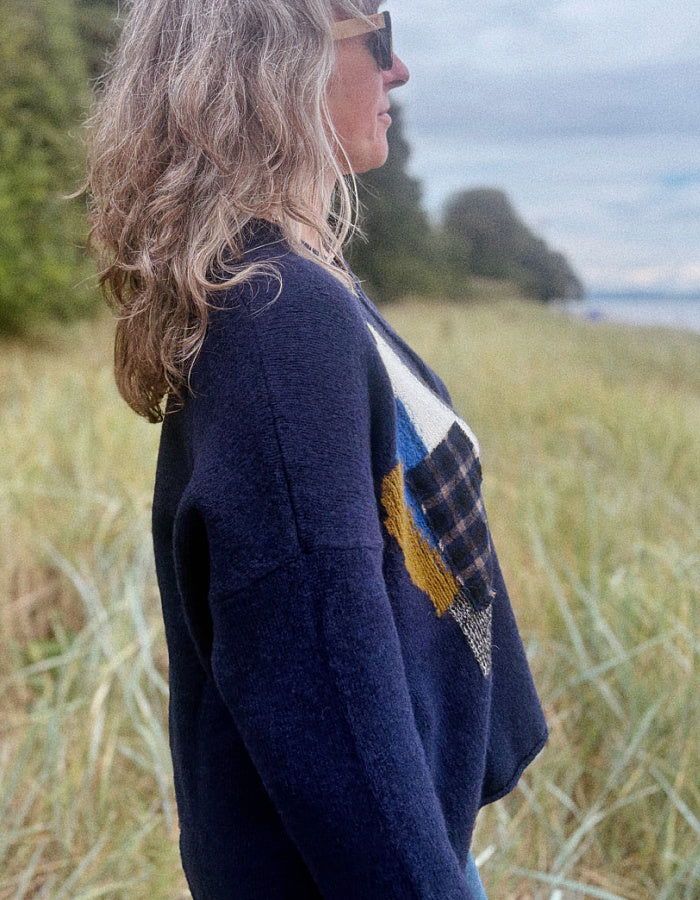 A woman in profile wearing a navy blue knit sweater with a multicolor patchwork design on the front, standing in a grassy outdoor field.