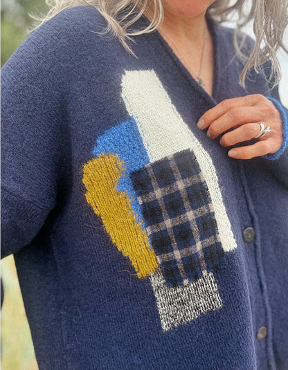 A close-up of a person wearing a navy blue knit cardigan with a large multicolor abstract patchwork design on the front, including cream, blue, mustard yellow, and plaid sections.