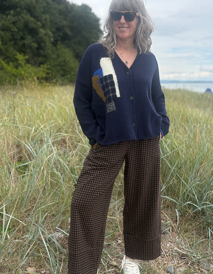 A woman wearing a navy blue knit cardigan with a multicolor geometric patchwork design on the chest, paired with brown and black checkered wide-leg pants, standing outdoors in a grassy field.