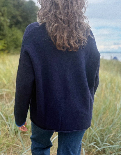 A person viewed from behind wearing an oversized dark navy knit sweater and blue jeans, standing in a grassy outdoor area with trees and water in the background.