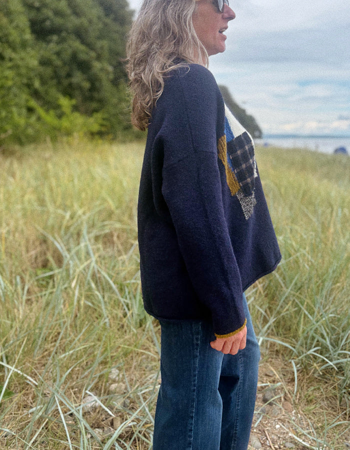 A woman wearing a dark navy knit sweater with a patchwork design on the front, standing outdoors in a grassy field with trees and water in the background.