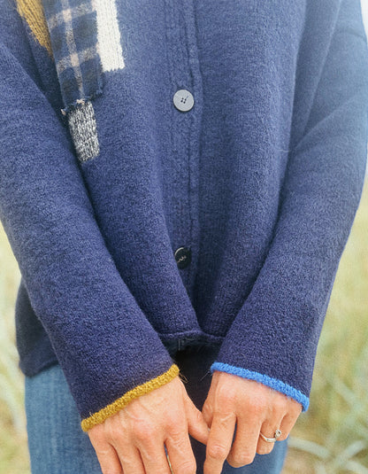 Close-up of a person wearing a navy blue knit cardigan with contrasting mustard yellow and light blue ribbed cuffs, paired with blue jeans.