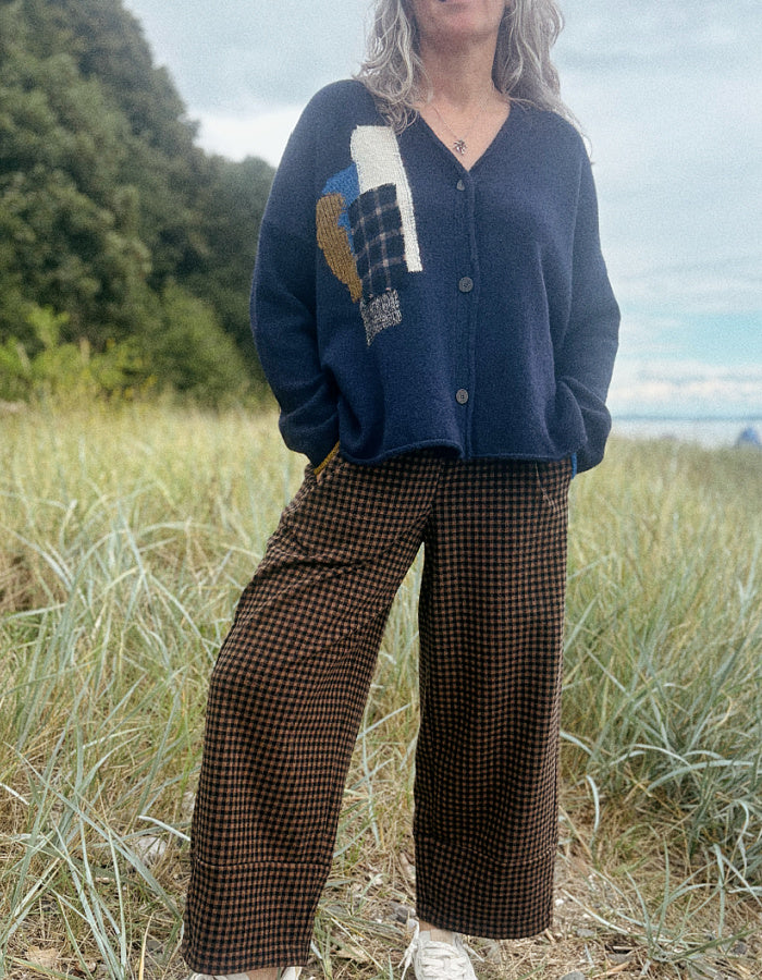A woman standing outdoors in a grassy field wearing a navy blue knit cardigan with a multicolor patchwork design on the front and brown wide-leg houndstooth pants.
