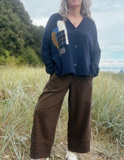 A woman standing outdoors in a grassy field wearing a navy blue knit cardigan with a multicolor patchwork design on the front and brown wide-leg houndstooth pants.