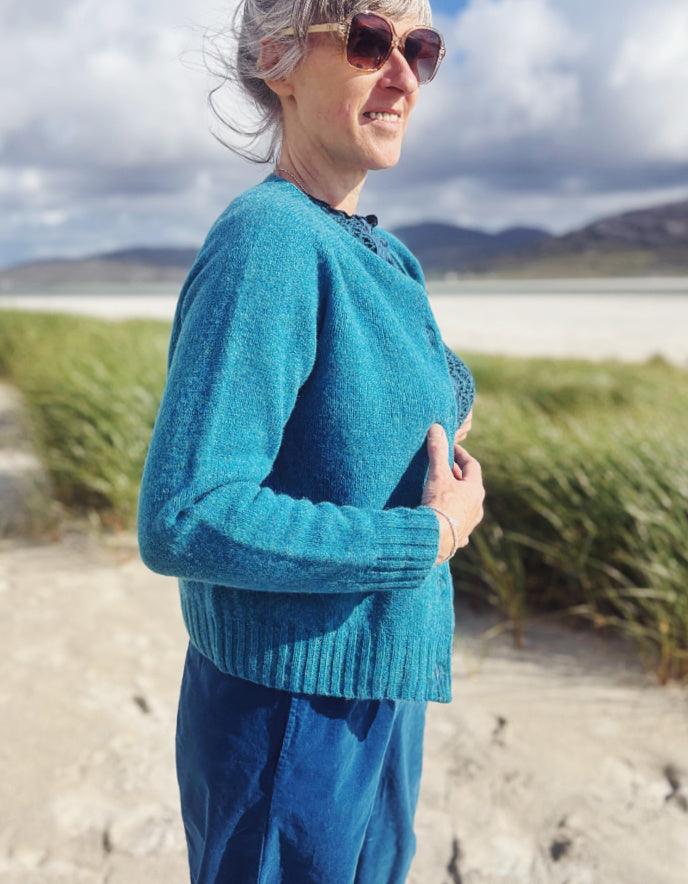A woman wearing a teal blue knit cardigan and matching blue pants stands outdoors on a sandy path with grass, water, and mountains in the background.