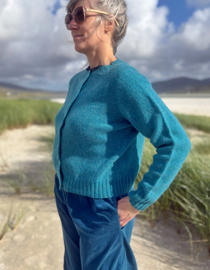 A woman wearing a teal blue knit cardigan and matching blue pants stands outdoors on a sandy path with beach grass, mountains, and a cloudy sky in the background.
