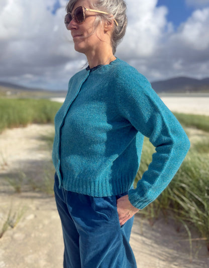 A woman wearing a teal blue knit cardigan and matching blue pants stands outdoors on a sandy path with beach grass, mountains, and a cloudy sky in the background.