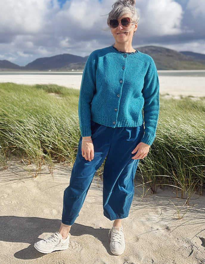 A woman standing on a sandy beach wearing a teal blue knit cardigan, matching blue jogger pants, and white sneakers, with dune grass and mountains in the background.