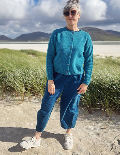A woman standing on a sandy beach wearing a teal blue knit cardigan, matching blue jogger pants, and white sneakers, with dune grass and mountains in the background.