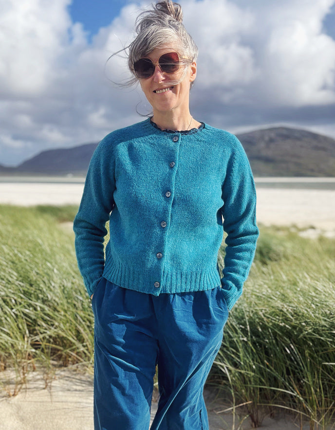 A woman wearing a teal blue button-up knit cardigan and matching blue pants stands on a beach dune with tall grass and mountains in the background.