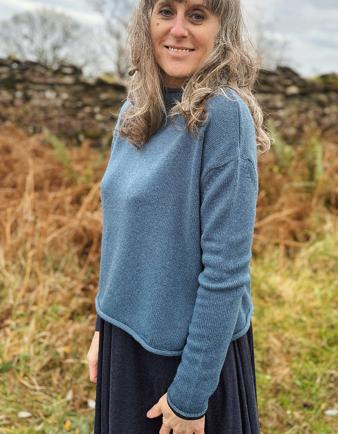 Woman wearing a blue sweater standing in a natural setting with dry grass and stone walls.
