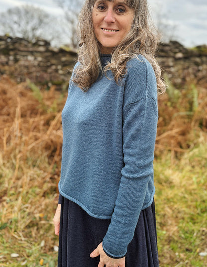 Woman wearing a blue sweater standing in a natural setting with dry grass and stone walls.
