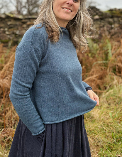 Woman wearing a blue sweater and dark skirt standing in a natural setting with dry grass and stone wall.