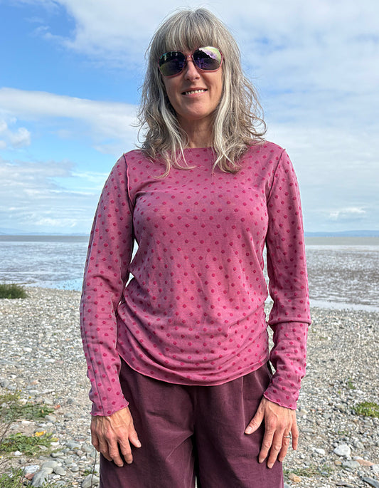 Woman wearing a pink long-sleeve top and purple pants standing on a beach with blue sky and water in the background.