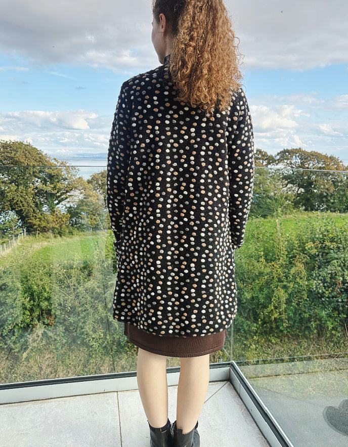 Person wearing a black polka dot dress standing in front of a window with a view of greenery.