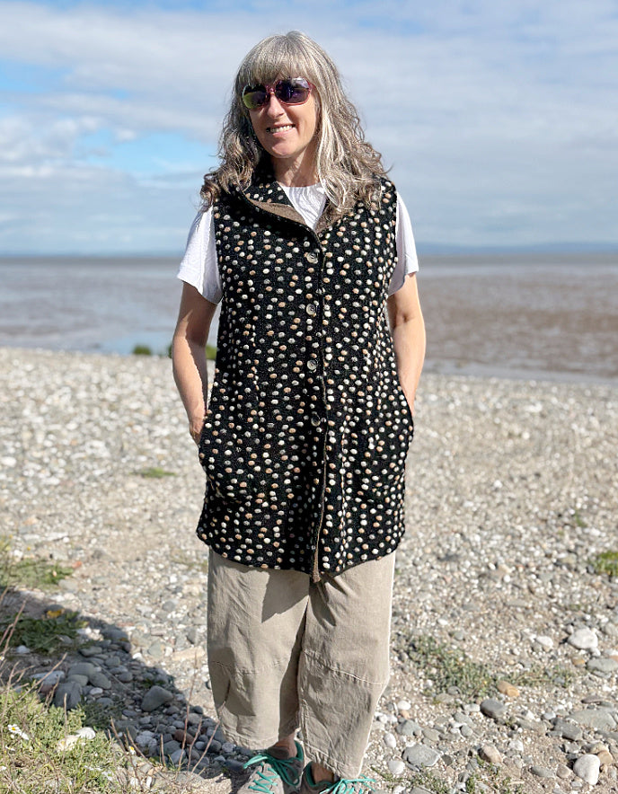 Woman wearing a black polka dot vest standing on a pebbly beach with water and sky in the background.