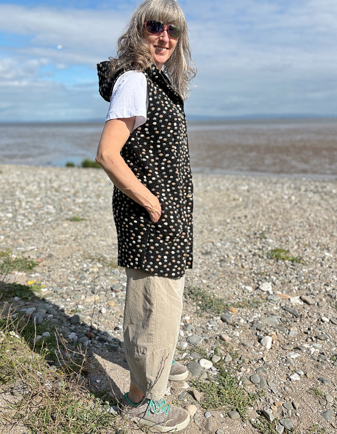 Person standing on a pebbly beach wearing a black polka dot dress and sunglasses.